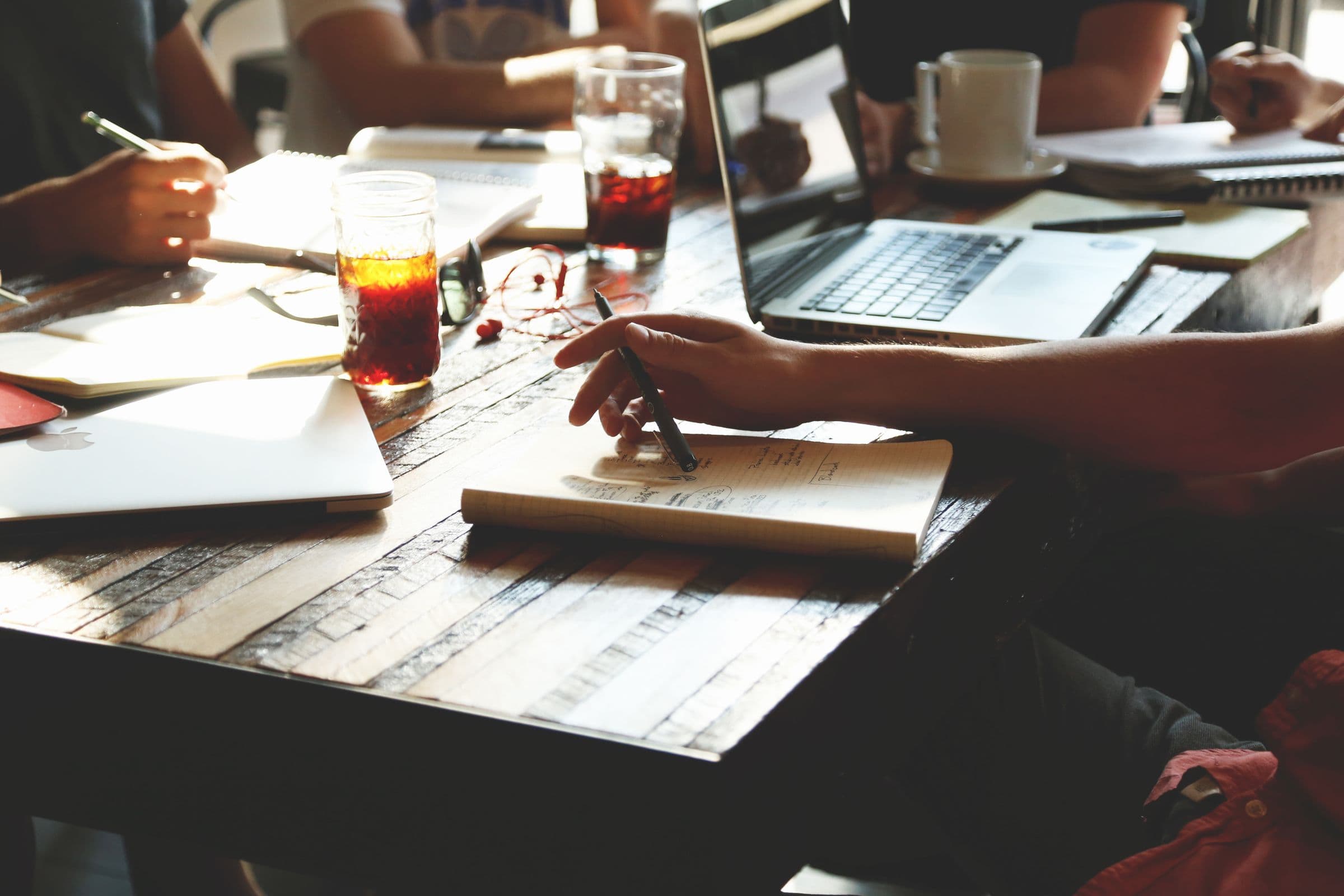 Hand die in een notitieboek schrijft aan een houten tafel met een laptop en glazen
